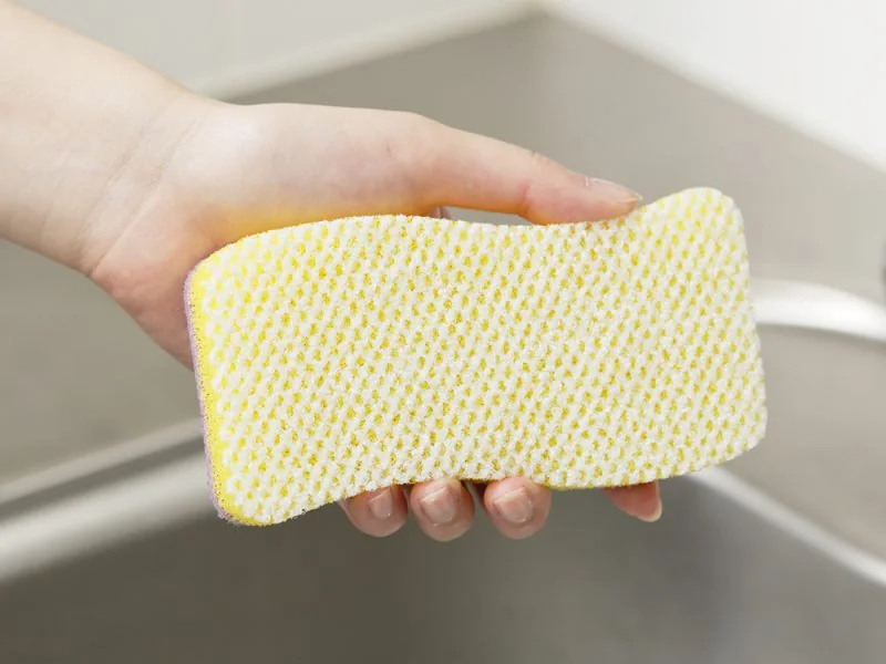 A hand holding a yellow kitchen sponge with a scrub layer